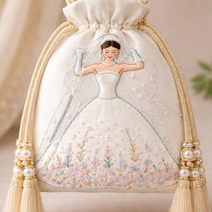 Peut inclure: Sac à cordon ivoire avec un motif de mariée brodé. Le sac présente une mariée en robe bustier avec un voile, les bras levés. La conception comprend une broderie florale aux couleurs pastel, des perles et des détails de glands.