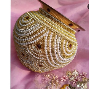 May include: A golden pot decorated with white pearls and red jewels. The pot is a traditional Indian ceremonial item.