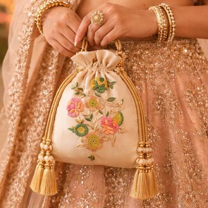 Peut inclure: Sac à cordon crème avec broderie florale rose, verte et jaune. Le sac est orné de poignées et de pompons dorés. En arrière-plan, un vêtement pêche avec des ornements dorés.