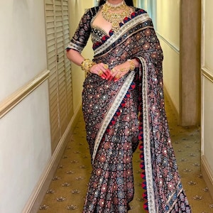 Puede incluir: Una mujer con un sari indio tradicional con un fondo negro y un patrón geométrico en rojo, azul y blanco. El sari tiene un borde decorativo y se combina con joyas de oro.