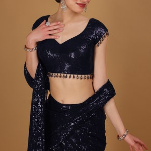 May include: A navy blue sequined saree with a matching sequined blouse. The blouse has a V-neckline and short sleeves with beaded embellishments. The saree is draped around the model's body and has a beaded border.