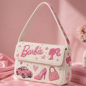 May include: A white beaded shoulder bag featuring pink beaded designs including the Barbie logo, a silhouette profile, a convertible car, a high-heeled shoe, a handbag, hearts, and stars. The strap is also decorated with pink beaded stars and hearts.