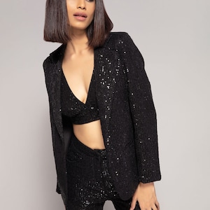 Puede incluir: Un blazer negro de lentejuelas, pantalones y top corto. El blazer tiene cuello en V y cierre de un solo botón. Los pantalones son ajustados y tienen cintura alta. El top corto es sin mangas y tiene cuello en V.