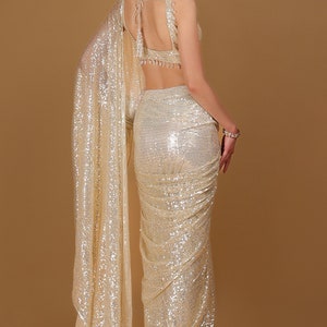 May include: A gold sequined saree with a matching sleeveless blouse. The saree is draped in a traditional style and has a shimmering, iridescent finish.
