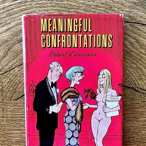 Puede incluir: Un libro vintage titulado "Meaningful Confrontations" de Osbert Lancaster. La portada presenta una ilustración de dibujos animados de tres personas frente a un fondo rojo. El libro está sobre una superficie de madera.