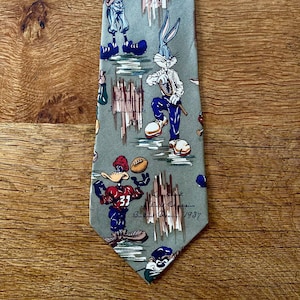 Può includere: Una cravatta originale con sfondo verde chiaro raffigurante personaggi dei cartoni animati che praticano sport. La cravatta presenta immagini di Bugs Bunny, Daffy Duck e Wile E. Coyote in abbigliamento da baseball, tennis e football americano. Il testo "Burbank Staggers 1937" è presente.