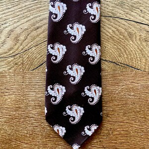 May include: A dark brown tie featuring a repeating paisley pattern in white and orange. The tie is made of a smooth fabric and is displayed on a wooden surface. The paisley design is detailed, adding a classic touch to the tie.