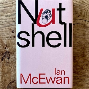 May include: A book cover for "Nutshell" by Ian McEwan. The cover is light pink with the title in large black letters and the author's name in red. The "U" in "Nutshell" is red and features a small illustration.