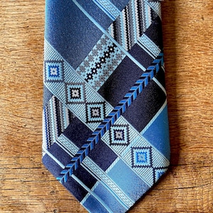 May include: A blue patterned tie with geometric designs in various shades of blue, including navy, light blue, and teal. The tie features diagonal stripes, squares, and zigzags, creating a visually interesting pattern. The tie is made of a smooth, possibly silk, material.