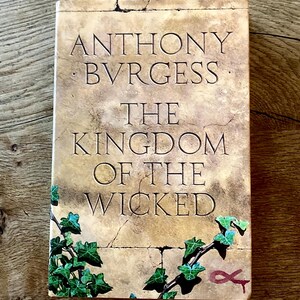 Könnte beinhalten: Ein Hardcover-Buch mit dem Titel "The Kingdom of the Wicked" von Anthony Burgess. Das Cover hat eine steinartige Textur mit eingraviertem Text und Efeu-Akzenten. Das Buch liegt auf einer Holzoberfläche.