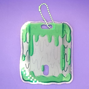 May include: A clear, rectangular ID badge holder with a green slime design around the edges. The holder has a small hole at the top with a green beaded chain attached. The background is a solid purple.