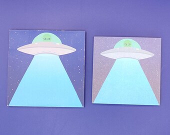 May include: Two square art prints featuring a white UFO with a green alien and a blue light beam. The background is a gradient of dark blue to light blue with white stars. The prints are displayed on a solid blue background.