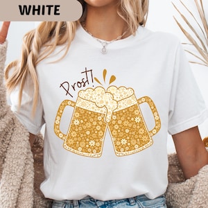 Könnte beinhalten: Weißes T-Shirt mit einem goldenen Motiv von zwei anstoßenden Bierkrügen, gefüllt mit Schaum. Das Design zeigt Blumenmuster und den Schriftzug "Prost!". Perfekt für Bierliebhaber und festliche Anlässe.