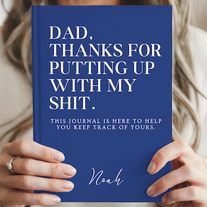 Könnte beinhalten: Ein blaues Notizbuch mit festem Einband und weißem Text, der lautet: DAD, THANKS FOR PUTTING UP WITH MY SHIT. THIS JOURNAL IS HERE TO HELP YOU KEEP TRACK OF YOURS. Der Name Noah ist in einer weißen Schreibschrift unten auf dem Einband gedruckt.