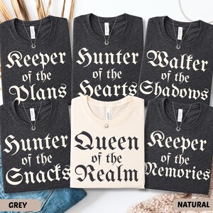 May include: A collection of dark grey and natural-colored t-shirts with various phrases in a gothic font. The phrases include "Keeper of the Plans," "Hunter of the Hearts," and "Queen of the Realm." Each shirt has a silver necklace.