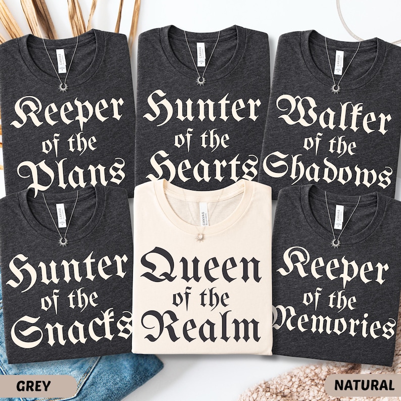 May include: A collection of dark grey and natural-colored t-shirts with various phrases in a gothic font. The phrases include "Keeper of the Plans," "Hunter of the Hearts," and "Queen of the Realm." Each shirt has a silver necklace.