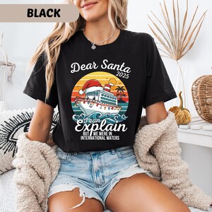 May include: Black t-shirt with a graphic featuring a cruise ship wearing a Santa hat, the text "Dear Santa 2025, I can explain, but we were in international waters." The design includes a sunset, palm trees, and waves.