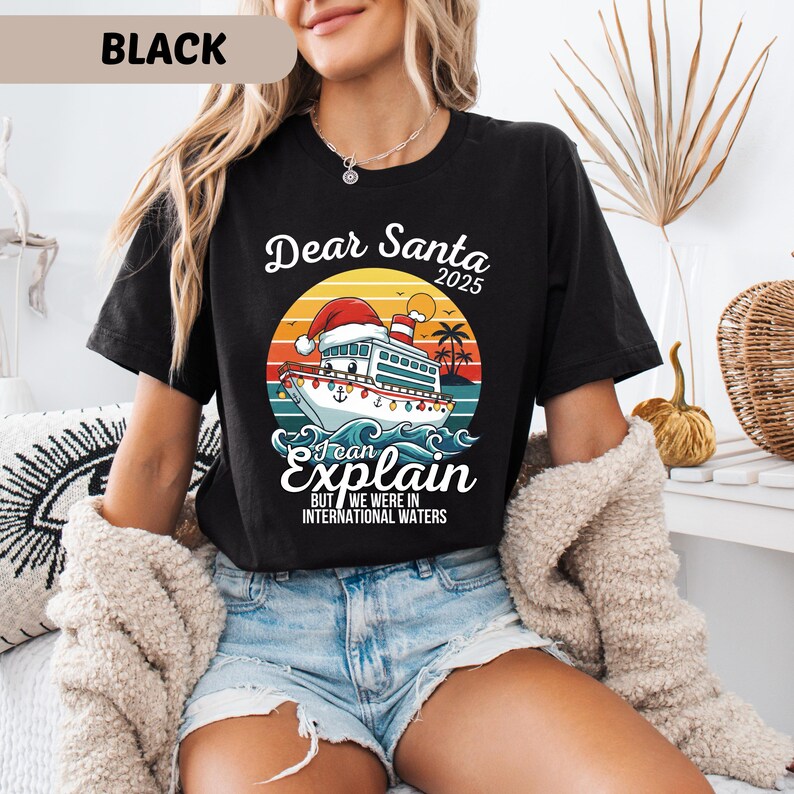 May include: Black t-shirt with a graphic featuring a cruise ship wearing a Santa hat, the text "Dear Santa 2025, I can explain, but we were in international waters." The design includes a sunset, palm trees, and waves.