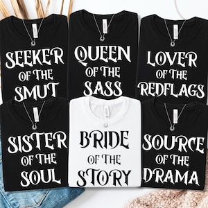 Könnte beinhalten: Eine Auswahl an schwarzen und weißen T-Shirts mit verschiedenen Sätzen in fetter weißer Schrift. Die Sätze lauten: "SEEKER OF THE SMUT", "QUEEN OF THE SASS", "LOVER OF THE REDFLAGS", "SISTER OF THE SOUL", "BRIDE OF THE STORY" und "SOURCE OF THE DRAMA". Jedes Shirt hat eine Halskette.