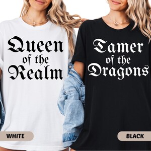 May include: Two t-shirts, one white and one black, with fantasy-themed text. The white shirt reads "Queen of the Realm" in a gothic font. The black shirt says "Tamer of the Dragons" in a similar style. The word "WHITE" is printed on the bottom of the white shirt, and "BLACK" on the black shirt.