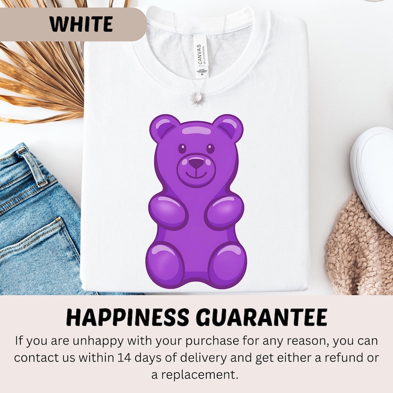 Puede incluir: Camiseta blanca con un dise&ntilde;o de osito de goma morado. La camiseta tiene la palabra "WHITE" en la parte superior y el texto "HAPPINESS GUARANTEE" en la parte inferior. La camiseta est&aacute; doblada y colocada sobre una superficie con vaqueros azules y un art&iacute;culo beige.