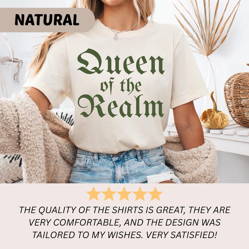 May include: Cream-colored t-shirt with the text "Queen of the Realm" in green gothic font. The word "NATURAL" is printed on a tan banner in the upper left corner. The shirt is paired with blue jeans.
