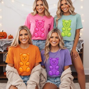 Puede incluir: Cuatro mujeres con camisetas coloridas con dise&ntilde;os de ositos de goma. Las camisetas son rosas, verdes, naranjas y moradas. El gr&aacute;fico del osito de goma est&aacute; centrado en la parte delantera de cada camiseta. Las mujeres sonr&iacute;en y llevan pantalones cortos vaqueros.