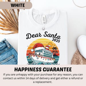 May include: White t-shirt with a graphic of a cruise ship wearing a Santa hat, the text "Dear Santa 2025", and a sunset background. The shirt is folded, with a pair of blue jeans and a white shoe visible.