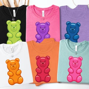 May include: A collection of six t-shirts in various colors, each featuring a gummy bear graphic. The shirts are black, pink, purple, white, orange, and light blue. Each shirt has a silver necklace with a sun pendant.