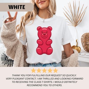 Puede incluir: Camiseta blanca con un dise&ntilde;o de osito de goma rojo. La palabra "WHITE" se muestra en la parte superior. La camiseta la lleva una persona. La imagen tambi&eacute;n incluye comentarios de clientes con cinco estrellas doradas.
