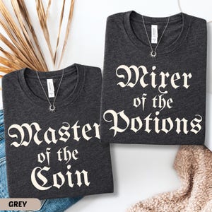 May include: Two dark grey t-shirts with white text that reads "Master of the Coin" and "Wirer of the Potions". Each shirt has a silver necklace with a sunburst pendant. The word "GREY" is at the bottom left.