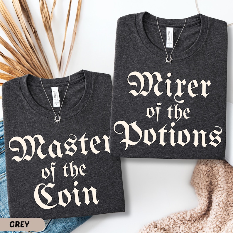 May include: Two dark grey t-shirts with white text that reads "Master of the Coin" and "Wirer of the Potions". Each shirt has a silver necklace with a sunburst pendant. The word "GREY" is at the bottom left.