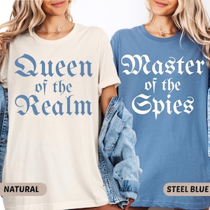 May include: Two t-shirts, one natural and one steel blue, each with a different medieval-style text design. The natural shirt reads "Queen of the Realm" in blue, while the steel blue shirt says "Master of the Spies" in white. Both shirts are short-sleeved.