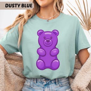 Puede incluir: Camiseta azul polvoriento con un dise&ntilde;o de oso de goma morado. La camiseta tiene mangas cortas y cuello redondo. El texto "DUSTY BLUE" est&aacute; en un rect&aacute;ngulo en la esquina superior izquierda.