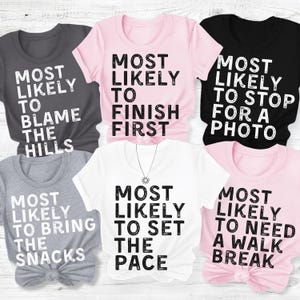 May include: A collection of six short-sleeved t-shirts in charcoal, light pink, black, heather gray, white, and light pink. Each shirt features bold, distressed black or white text stating phrases like Most Likely To Blame The Hills, Finish First, Stop For A Photo, Bring The Snacks, Set The Pace, and Need A Walk Break.