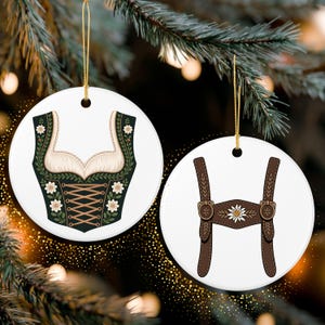 May include: Two round white Christmas ornaments with gold hanging strings. One ornament features a green and cream dirndl bodice design with floral accents. The other ornament displays brown lederhosen suspenders with a floral detail.
