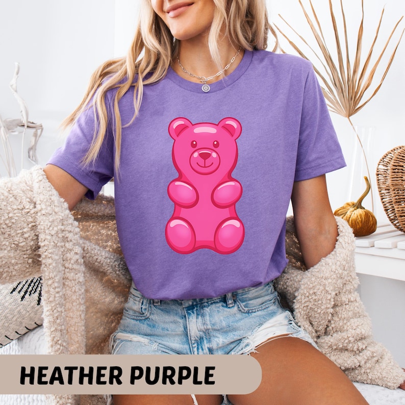 Puede incluir: Camiseta morada jaspeada con un gran dise&ntilde;o de osito de goma rosa brillante. El osito de goma tiene una cara sonriente y est&aacute; centrado en la camiseta. El texto "HEATHER PURPLE" se muestra en la parte inferior de la imagen.