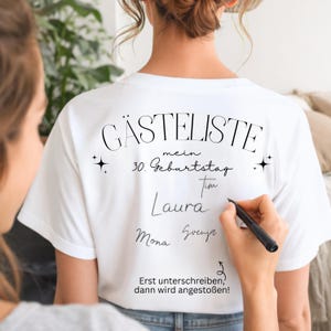 Könnte beinhalten: Weißes T-Shirt mit schwarzem Text und Unterschriften. Der Text auf dem Shirt lautet "GÄSTELISTE mein 30. Geburtstag" und "Erst unterschreiben, dann wird angestoßen!". Eine Person unterschreibt das Shirt mit einem schwarzen Stift.