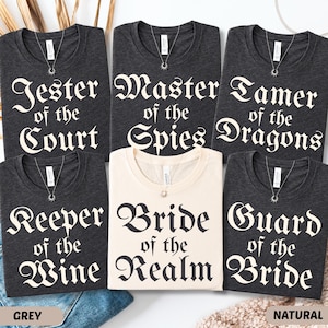 May include: A collection of t-shirts in grey and natural colors, each with a unique title in a gothic font. Titles include "Jester of the Court", "Master of the Spies", "Tamer of the Dragons", "Keeper of the Wine", "Bride of the Realm", and "Guard of the Bride".