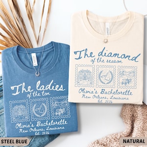 May include: Two t-shirts, one steel blue and one natural, with the text "The ladies of the ton" and "The diamond of the season." Each shirt features three postage stamp-style illustrations and the text "Olivia's Bachelorette, New Orleans, Louisiana, Est. 2026."