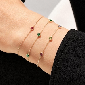 May include: Three delicate gold chain bracelets, each featuring small, round, bezel-set gemstones in various colors including red, green, and blue. The bracelets are worn together on the wrist, showcasing a fine link design with evenly spaced stones.
