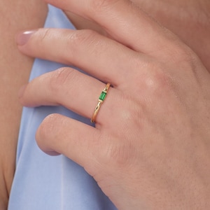 May include: A thin gold ring featuring a single, small, rectangular-cut green gemstone set horizontally in the center. The band is slender and polished, showcasing a minimalist design that highlights the vibrant emerald-colored stone.