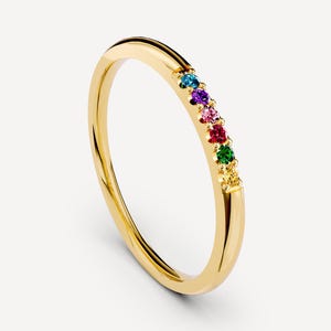 May include: A thin gold band ring featuring a row of six small, round, multi-colored gemstones set along the top. The stones are arranged in a sequence of blue, purple, pink, red, green, and yellow, creating a vibrant, rainbow-like accent.