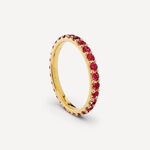 May include: A yellow gold eternity band featuring a continuous row of round-cut, vibrant red gemstones set in a shared-prong style. The ring has a polished metallic finish and a slim, circular profile, showcasing the stones around the entire band.