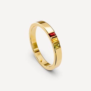 May include: A polished gold band ring featuring three vertically aligned baguette-cut gemstones in a channel setting. The stones are arranged in a gradient sequence, displaying a deep red, a bright yellow, and a light green hue respectively.