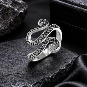 Könnte beinhalten: Ein silberfarbener Ring in Form eines gewundenen Tintenfischtentakels. Das Design zeichnet sich durch eine polierte metallische Oberfläche an den Außenkanten und eine dunkle, vertiefte Textur mit kreisförmigen Saugnapfdetails an der Innenkurve aus.
