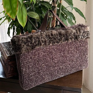May include: A rectangular, knitted tote bag in a muted purple hue, trimmed with faux fur in a leopard print. The bag features a brown handle and sits on a dark wooden surface, with lush green plants in the background.