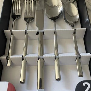 May include: A set of five silver-toned cutlery pieces in a white box with a black border. The set includes a knife, two forks, and two spoons. The box has the number "2" and the word "piece" printed on it.