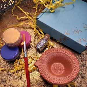 May include: A small terracotta diya, a traditional Indian oil lamp, sits on a gold surface surrounded by yellow tissue paper.  There are also three small jars of paint, a bottle of glitter, and a red paintbrush.