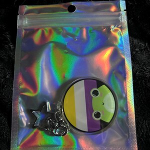 May include: A clear, iridescent zip-lock bag containing a keychain. The keychain features a round charm with a non-binary pride flag design and a green alien face. A silver star-shaped charm is also attached.